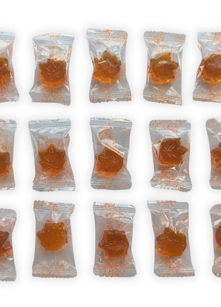 Fifteen individually wrapped Honey, Maple & Lemon hard candies arranged in three neat rows on a white background.  The image provides a clear view of the approximate quantity included in the 90 g package while showing the amber maple-leaf candies and their individual clear wrappers printed with orange maple-leaf outlines.