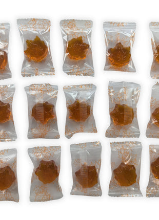 Fifteen individually wrapped maple-leaf hard candies arranged in three neat rows on a white background.  Each golden amber candy is visible through a clear wrapper decorated with orange maple-leaf outlines, giving customers a clear view of the approximate quantity, individual packaging, shape, and appearance of the candies inside the 90 g bag.