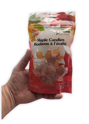 A hand holding the 90 g resealable package of maple hard candies against a clean white background to provide an accurate sense of the package’s size and proportions.  The bilingual front label, colourful maple-leaf design, transparent product window, and individually wrapped amber candies are all visible.