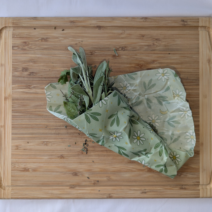 Green floral beeswax wrap cradling fresh herbs on a bamboo cutting board to show how it keeps leafy greens crisp