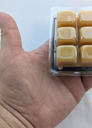 A hand holding a clear clamshell tray of six natural beeswax melt cubes on a white background, showing the size and shape of the wax pieces up close.  This image is useful for demonstrating scale and helping customers understand the cube size and overall product format.
