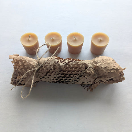 Four natural beeswax votive refill candles arranged above their kraft honeycomb paper wrapping and twine on a clean white background.  The image shows the full four-candle set, warm honey-gold wax colour, visible cotton wicks, and simple plastic-free presentation.
