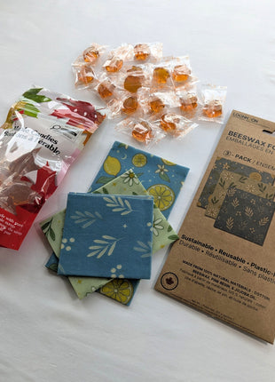 The Beeswax Wraps & Maple Candy Starter Set is arranged on a clean white background with a 3-pack of reusable beeswax food wraps, a 90 g bag of individually wrapped maple candies, loose candies, and all three folded wrap patterns.  This flat-lay image clearly shows the complete bundle and the products included.