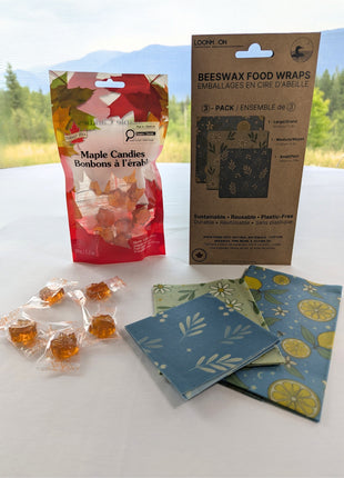 Reusable beeswax food wraps and maple candies are displayed together with both packages upright against a softly blurred British Columbia mountain landscape.  The three folded wrap sizes and several individually wrapped candies are visible in the foreground, giving the starter set a natural Canadian lifestyle presentation.