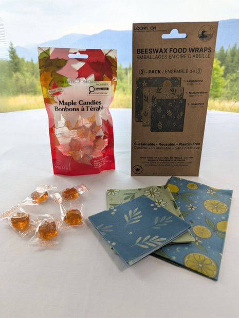 Reusable beeswax food wraps and maple candies are displayed together with both packages upright against a softly blurred British Columbia mountain landscape.  The three folded wrap sizes and several individually wrapped candies are visible in the foreground, giving the starter set a natural Canadian lifestyle presentation.