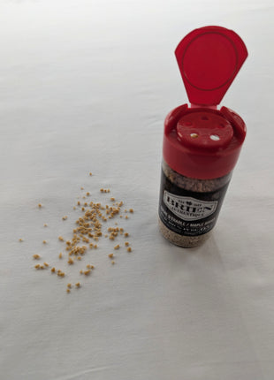 This image shows a Brien Authentique maple sugar shaker with the red flip-top lid open beside a small spill of coarse maple sugar crystals on a light surface.  The shaker stands upright with the front label partially visible, helping illustrate how the product is dispensed and used.  This lifestyle-style image is helpful for showing the texture of the maple sugar and how it can be sprinkled onto foods and drinks.