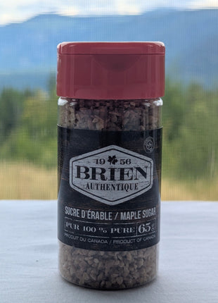 This image features a Brien Authentique maple sugar shaker photographed in front of a scenic mountain and forest background.  The shaker has a red lid, a dark rustic label, and visible maple sugar crystals inside.  The front label identifies the product as pure maple sugar, 65 g, and Product of Canada.  This lifestyle image adds a natural Canadian feel and helps show the product in a more atmospheric setting.