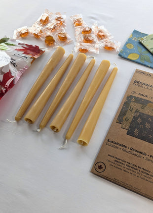 An angled product view shows the Canadian Maple and Beeswax Kitchen Set with six beeswax taper candles, three folded reusable food wraps, and individually wrapped maple candies beside their retail packages.  The perspective highlights the length and natural wax colour of the tapers while giving a clear view of the wrap patterns and maple treat assortment.