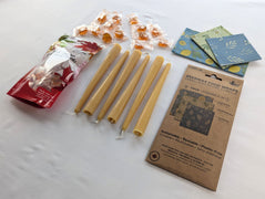 An angled product view shows the Canadian Maple and Beeswax Kitchen Set with six beeswax taper candles, three folded reusable food wraps, and individually wrapped maple candies beside their retail packages.  The perspective highlights the length and natural wax colour of the tapers while giving a clear view of the wrap patterns and maple treat assortment.