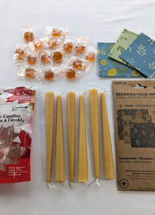 The Canadian Maple and Beeswax Kitchen Set is arranged in a clean overhead flat lay with six natural beeswax taper candles, a 3-pack of reusable beeswax food wraps, and a 90 g bag of individually wrapped maple candies.  The image clearly shows the complete bundle, including the three wrap patterns, both retail packages, and approximately fifteen amber maple candies.