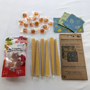 The Canadian Maple and Beeswax Kitchen Set is arranged in a clean overhead flat lay with six natural beeswax taper candles, a 3-pack of reusable beeswax food wraps, and a 90 g bag of individually wrapped maple candies.  The image clearly shows the complete bundle, including the three wrap patterns, both retail packages, and approximately fifteen amber maple candies.