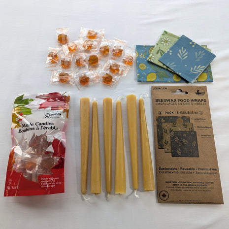The Canadian Maple and Beeswax Kitchen Set is arranged in a clean overhead flat lay with six natural beeswax taper candles, a 3-pack of reusable beeswax food wraps, and a 90 g bag of individually wrapped maple candies.  The image clearly shows the complete bundle, including the three wrap patterns, both retail packages, and approximately fifteen amber maple candies.