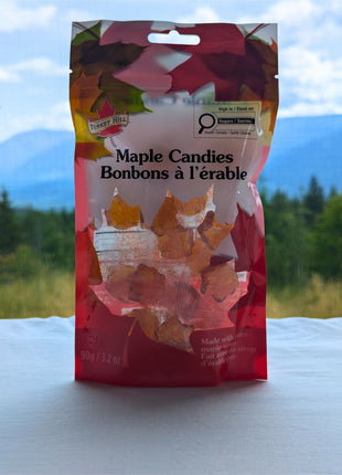A 90 g package of maple hard candies displayed in front of a scenic British Columbia landscape with forest, open grassland, distant blue mountains, and a bright cloud-filled sky.  The bilingual package and wrapped maple-leaf candies remain visible while the natural setting adds an authentic Canadian outdoor context.