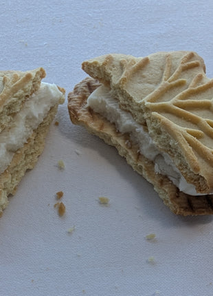 Broken Canadian maple cream sandwich cookie showing the thick cream filling between two maple-leaf-shaped cookies. This close-up reveals the crisp baked cookie texture and creamy centre of a Canadian maple cookie made with pure maple syrup.