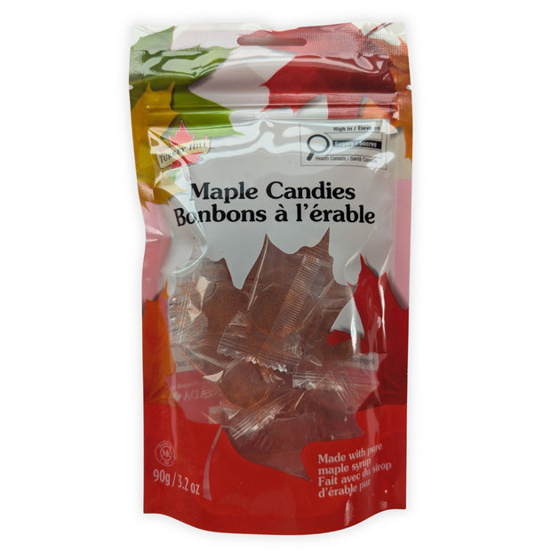 A front view of a 90 g resealable package of maple hard candies made with pure maple syrup, photographed on a clean white background.  The bilingual Maple Candies and Bonbons à l’érable label is clearly visible, along with the red maple-leaf packaging and individually wrapped amber maple-leaf candies shown through the transparent window.