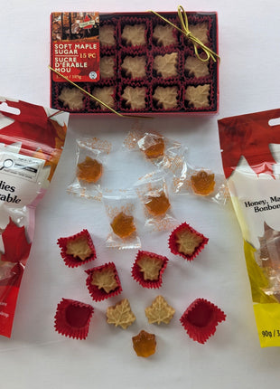 Overhead view of the Canadian maple treat sampler with all three packages surrounded by wrapped and unwrapped maple candies.  The image shows the different textures, shapes, colours, and presentation of the soft maple sugar leaves, traditional hard maple candies, and honey, maple and lemon candies included in the assortment.