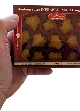 A 100 g box of Canadian maple sugar candies held in one hand to show the size and scale of the package. Maple-leaf-shaped candies are visible through the window and are made with pure maple syrup.