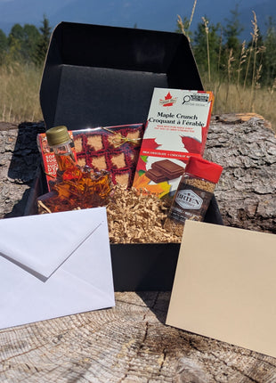 LOONMOON Core Maple Gift Box displayed outdoors against a British Columbia mountain backdrop with Canadian maple syrup in a leaf-shaped glass bottle, soft maple candies, maple sugar, and a maple milk chocolate bar arranged in a black gift box.  A blank folded greeting card and matching envelope are shown in front, highlighting the complete gift-ready presentation.