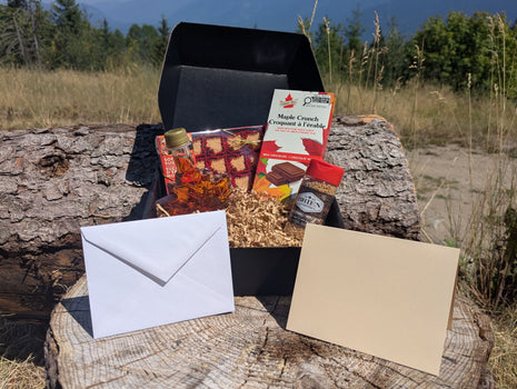 LOONMOON Core Maple Gift Box displayed outdoors against a British Columbia mountain backdrop with Canadian maple syrup in a leaf-shaped glass bottle, soft maple candies, maple sugar, and a maple milk chocolate bar arranged in a black gift box.  A blank folded greeting card and matching envelope are shown in front, highlighting the complete gift-ready presentation.