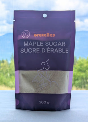A 200 g pouch of fine maple sugar displayed against a scenic British Columbia landscape with green forest, distant blue mountains and a cloud-filled sky.  The purple bilingual package and transparent product window remain visible while the outdoor setting adds natural Canadian context.