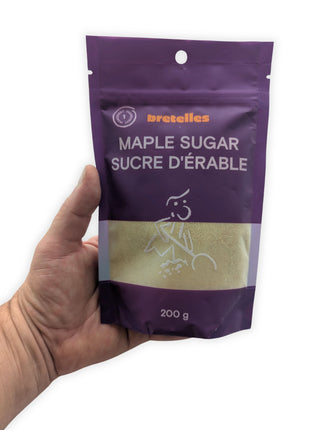 A hand holding the 200 g resealable pouch of fine maple sugar against a clean white background to show the package’s actual size and proportions.  The bilingual Maple Sugar and Sucre d’érable label, purple packaging and transparent window revealing the golden sugar are clearly visible.