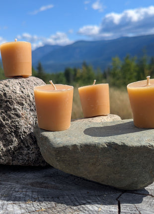 Four unwrapped beeswax votive candles arranged on natural stones over a weathered stump with forest, blue sky, clouds, and British Columbia mountains in the background.  The outdoor lifestyle image shows the full four-pack while emphasizing the candles’ natural wax colour and handmade character.