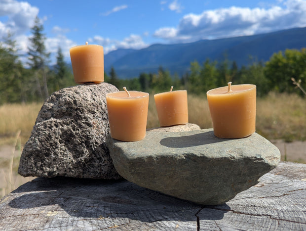 Four unwrapped beeswax votive candles arranged on natural stones over a weathered stump with forest, blue sky, clouds, and British Columbia mountains in the background.  The outdoor lifestyle image shows the full four-pack while emphasizing the candles’ natural wax colour and handmade character.