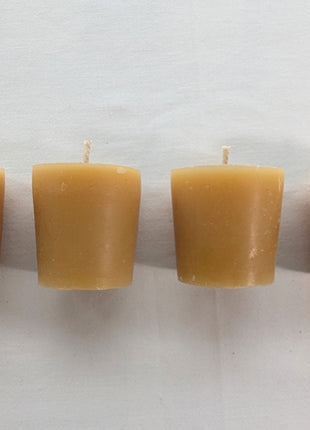 Four unwrapped beeswax votive refill candles are lined up against a clean white background.  The image clearly shows their compact tapered shape, warm natural wax colour, cotton wicks, and slight handmade variation.