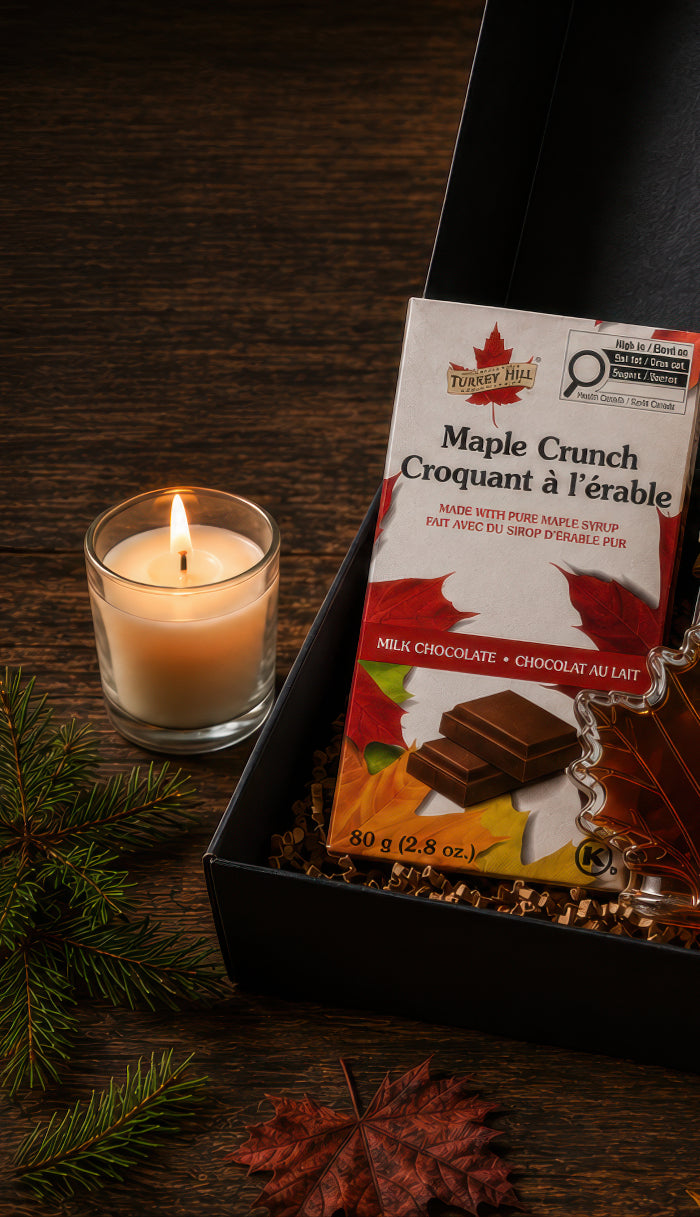 Open black Canadian maple gift box with maple syrup, maple sugar, maple chocolate, soft maple candies, and a lit beeswax candle on a dark rustic wood table.