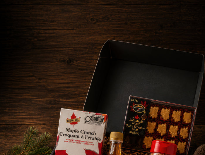 Open black Canadian maple gift box with maple syrup, maple sugar, maple chocolate, soft maple candies, and a lit beeswax candle on a dark rustic wood table.
