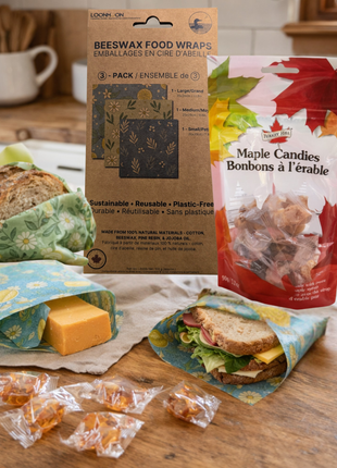 Beeswax food wraps and maple candies displayed on a rustic kitchen countertop, with bread, cheese, and a sandwich wrapped in reusable wraps beside packaged maple candy treats.