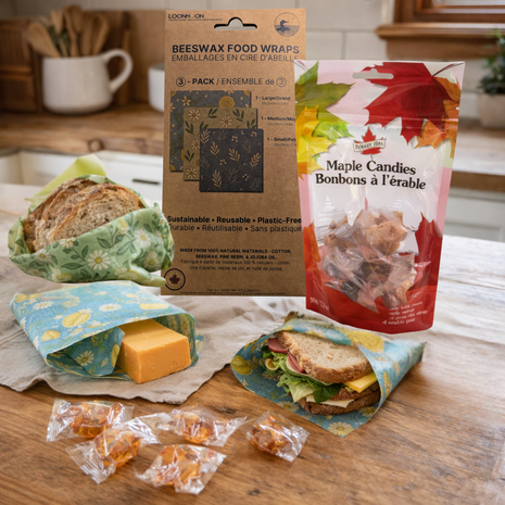 Beeswax food wraps and maple candies displayed on a rustic kitchen countertop, with bread, cheese, and a sandwich wrapped in reusable wraps beside packaged maple candy treats.