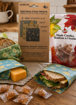 Beeswax food wraps and maple candies displayed on a rustic kitchen countertop, with bread, cheese, and a sandwich wrapped in reusable wraps beside packaged maple candy treats.