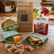 Beeswax food wraps and maple candies displayed on a rustic kitchen countertop, with bread, cheese, and a sandwich wrapped in reusable wraps beside packaged maple candy treats.