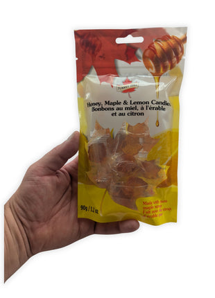A hand holding the 90 g resealable package of Honey, Maple & Lemon Candies against a clean white background to provide an accurate sense of the pouch’s size and proportions.  The bilingual label, honey and lemon artwork, transparent product window and individually wrapped maple-leaf candies are visible.