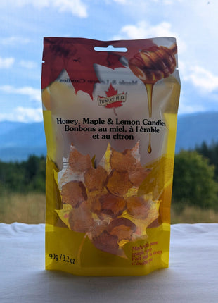 A 90 g package of Honey, Maple & Lemon Candies displayed against a scenic British Columbia landscape with forest, open grassland, distant blue mountains and a bright cloud-filled sky.  The bilingual package and individually wrapped maple-leaf candies remain clearly visible while the natural setting adds authentic Canadian outdoor context.