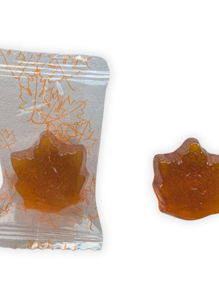 A close-up of one individually wrapped Honey, Maple & Lemon hard candy beside an unwrapped candy on a clean white background.  The image shows the clear wrapper decorated with orange maple leaves along with the candy’s detailed maple-leaf shape, golden amber colour, glossy surface and translucent appearance.