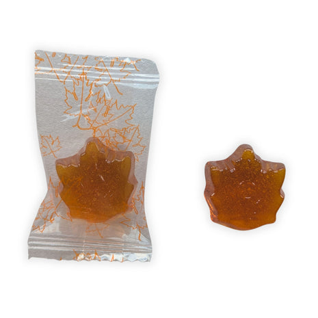 A close-up of one individually wrapped Honey, Maple & Lemon hard candy beside an unwrapped candy on a clean white background.  The image shows the clear wrapper decorated with orange maple leaves along with the candy’s detailed maple-leaf shape, golden amber colour, glossy surface and translucent appearance.