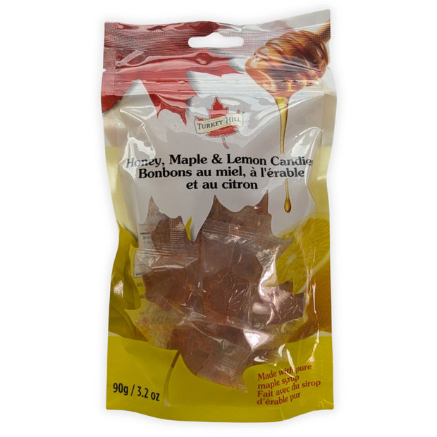 A front view of a 90 g resealable package of Honey, Maple & Lemon Candies photographed on a clean white background.  The bilingual English and French label, honey dipper and lemon artwork, colourful maple-leaf design, and individually wrapped amber maple-leaf hard candies are clearly visible through the transparent package window.