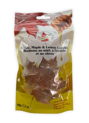 A front view of a 90 g resealable package of Honey, Maple & Lemon Candies photographed on a clean white background.  The bilingual English and French label, honey dipper and lemon artwork, colourful maple-leaf design, and individually wrapped amber maple-leaf hard candies are clearly visible through the transparent package window.
