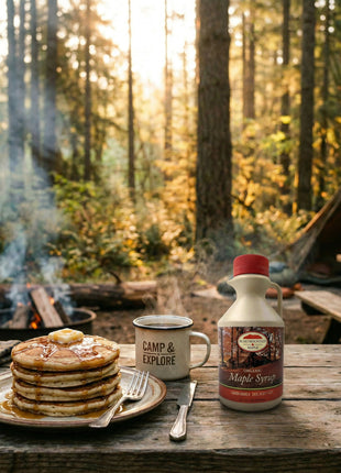 Small 500ml jug of organic maple syrup on a rustic picnic table beside pancakes and a camping mug, with a forest campsite and campfire in the background