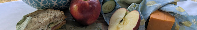 Overhead view of reusable beeswax wraps used to cover a bowl of cherries and wrap a sandwich, cucumber, cut onion, cheddar cheese, and fresh apple.  This practical food-storage arrangement shows how different wrap sizes can help keep produce, snacks, cheese, leftovers, and packed lunches covered and organized.