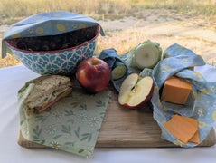 Overhead view of reusable beeswax wraps used to cover a bowl of cherries and wrap a sandwich, cucumber, cut onion, cheddar cheese, and fresh apple.  This practical food-storage arrangement shows how different wrap sizes can help keep produce, snacks, cheese, leftovers, and packed lunches covered and organized.