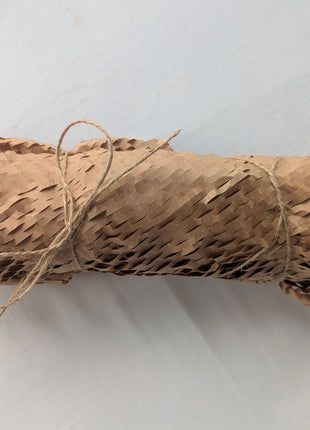 A six-candle beeswax taper set wrapped in expandable honeycomb kraft paper and secured with natural twine on a soft white background.  The photograph shows the protective, low-plastic presentation used to keep the candles together during storage and shipping.