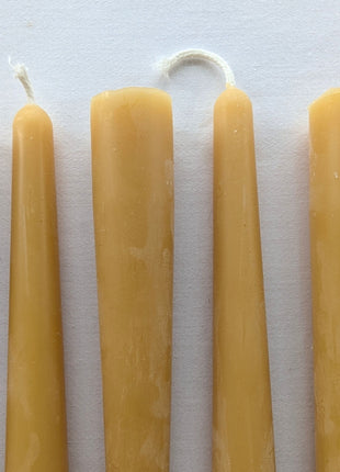 Close-up view of six natural beeswax taper candles on a white background, showing their rounded tops, cotton wicks and authentic wax surface texture.  The detailed photograph highlights the natural differences in wick shape, finish and golden beeswax colour across the six-candle set.