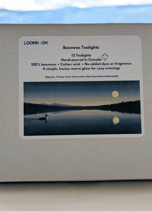 A closed LOONMOON box of twelve beeswax tealights is shown against a softly blurred British Columbia mountain background.  The front label identifies the hand-poured Canadian candles as 100% beeswax with cotton wicks, no added dyes or fragrance, and an approximate five-hour burn time per tealight when burned continuously.