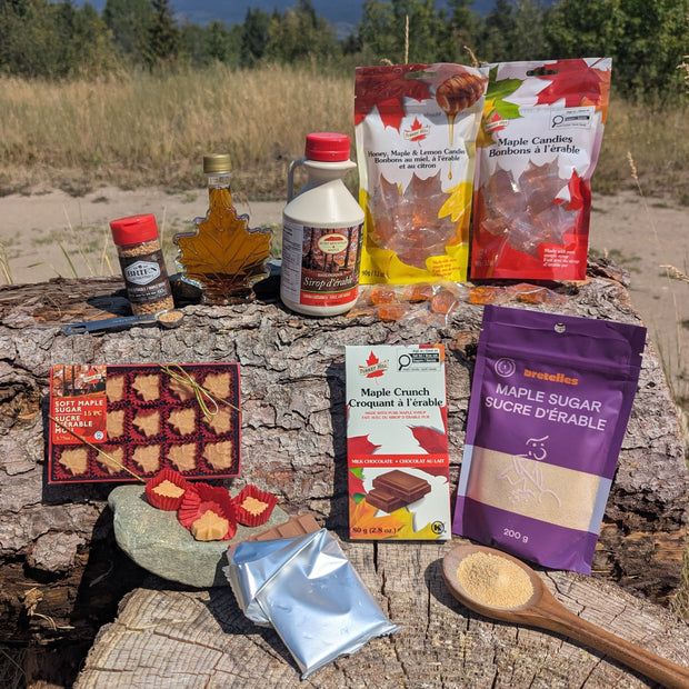 A collection of Canadian maple treats outdoors on rustic wood with a mountain landscape. Features pure maple syrup, maple sugar, maple chocolates, soft maple candies, hard maple candies, honey maple lemon candies, and maple sugar crystals in a natural forest setting that reflects the authentic Canadian origin of the products. The real photography highlights LOONMOON's premium collection while creating a warm, trustworthy shopping experience for customers looking for genuine Canadian maple gifts and treats.