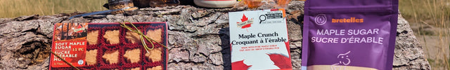 A collection of Canadian maple treats outdoors on rustic wood with a mountain landscape. Features pure maple syrup, maple sugar, maple chocolates, soft maple candies, hard maple candies, honey maple lemon candies, and maple sugar crystals in a natural forest setting that reflects the authentic Canadian origin of the products. The real photography highlights LOONMOON's premium collection while creating a warm, trustworthy shopping experience for customers looking for genuine Canadian maple gifts and treats.