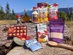 A collection of Canadian maple treats outdoors on rustic wood with a mountain landscape. Features pure maple syrup, maple sugar, maple chocolates, soft maple candies, hard maple candies, honey maple lemon candies, and maple sugar crystals in a natural forest setting that reflects the authentic Canadian origin of the products. The real photography highlights LOONMOON's premium collection while creating a warm, trustworthy shopping experience for customers looking for genuine Canadian maple gifts and treats.