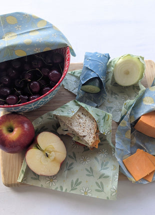 Reusable beeswax food wraps shown in everyday kitchen use with a covered bowl of cherries, wrapped cucumber and onion, cheddar cheese, a sandwich, and a cut red apple.  The natural wraps help cover bowls, protect produce, store cheese, and pack lunches while offering a practical reusable alternative to disposable plastic wrap.
